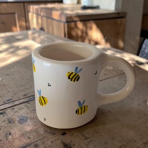 Può includere: Una tazza in ceramica color crema con un motivo dipinto a mano di piccole api gialle e nere con ali azzurre. La tazza presenta un manico spesso e arrotondato e una finitura liscia e lucida, con piccoli punti neri sparsi tra le api.