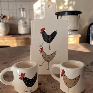 Può includere: Un tagliere da portata in ceramica bianca con un foro nella parte superiore e due tazze in ceramica bianca abbinate. Il tagliere presenta due polli dipinti, uno nero a pois bianchi e uno beige a pois neri. Ogni tazza mostra uno dei disegni.