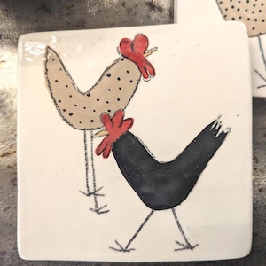 Op de afbeelding: Vierkante keramische tegel met een handgetekende illustratie van twee kippen. Eén kip is bruin met zwarte stippen, de andere is zwart. Beide hebben rode kammen tegen een witte achtergrond. De tegel heeft een glanzende afwerking.