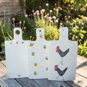 Può includere: Tre taglieri da portata rettangolari in ceramica bianca con manici. Il primo tagliere presenta piccoli cuori rossi. Il secondo mostra un motivo di api gialle e nere con ali blu. Il terzo tagliere raffigura due polli neri stilizzati.
