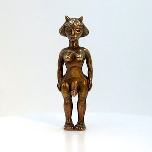 May include: A bronze-colored figurine of a nude figure with a stylized face and cat-like ears. The figure is standing upright with arms at its sides. The sculpture has a smooth, polished finish and is set against a white background.