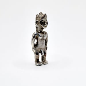 May include: A small, silver figurine of a humanoid figure with a crown-like headpiece. The figure is standing upright with arms at its sides. The figurine is on a white background.