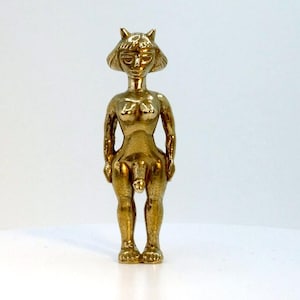 May include: A gold-colored figurine of a nude person with a cat-like head. The figure is standing upright with arms at its sides. The figurine has detailed facial features and is standing on a white surface.