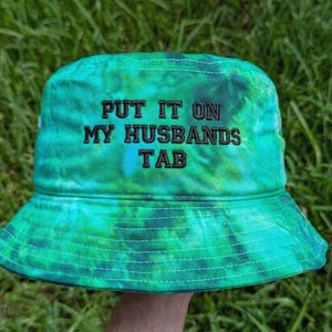 May include: A bucket hat featuring a vibrant green and blue tie-dye pattern. The front of the hat displays the embroidered black text that reads, "PUT IT ON MY HUSBANDS TAB." The hat has a wide, downward-sloping brim and visible stitching details.