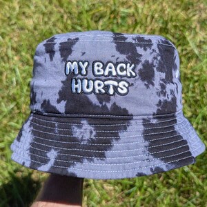 May include: A bucket hat featuring a grey and black tie-dye pattern. The front of the hat displays the embroidered text MY BACK HURTS in white, bubbly, outlined letters. The hat has a downward-sloping brim and visible stitching along the crown.