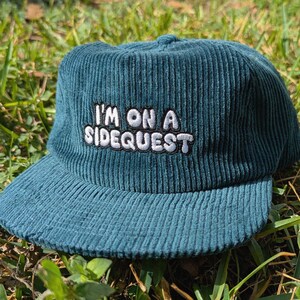 May include: Teal corduroy baseball cap with the embroidered text "I'M ON A SIDEQUEST" in white. The hat has a curved brim and a structured crown. The cap is sitting on a bed of green grass.