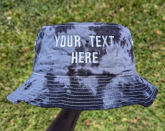 May include: A black and blue tie-dye bucket hat with the text "YOUR TEXT HERE" embroidered in white on the front.
