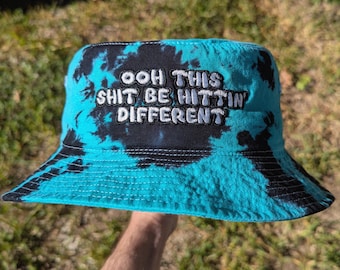 May include: A turquoise tie-dye bucket hat with the embroidered phrase "OOH THIS SHIT BE HITTIN' DIFFERENT" in white lettering. The hat features a black brim and a black and turquoise tie-dye pattern.