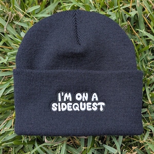 May include: A black knit beanie featuring a folded cuff and white embroidered text that reads I'M ON A SIDEQUEST. The cap is constructed from a ribbed fabric with a consistent dark tone and clear, bold lettering centered on the front panel.