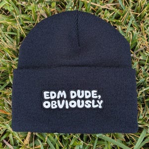 May include: A black beanie with the text "EDM DUDE, OBVIOUSLY" in white, block letters. The beanie is folded at the brim and sits on a bed of green grass. The hat is made of a knitted material.