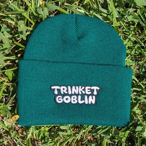 May include: A dark green beanie hat with the text "TRINKET GOBLIN" embroidered in white with a black outline. The hat is made of a knitted material and is displayed on a bed of green grass.