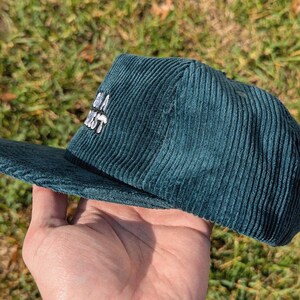 May include: Teal corduroy baseball cap with a flat brim. The cap features white embroidered text. The hat is held in a hand against a background of green grass.