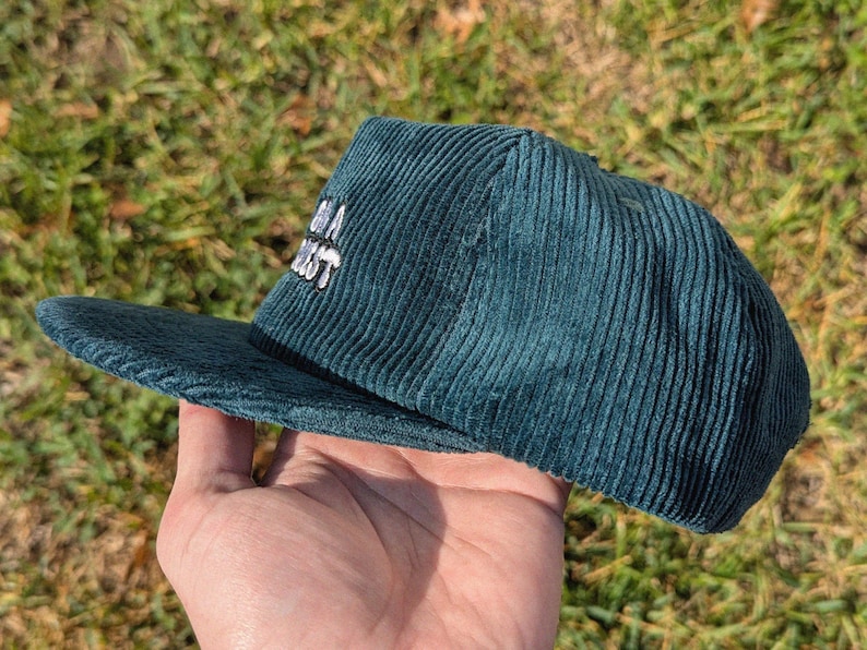 May include: Teal corduroy baseball cap with a flat brim. The cap features white embroidered text. The hat is held in a hand against a background of green grass.
