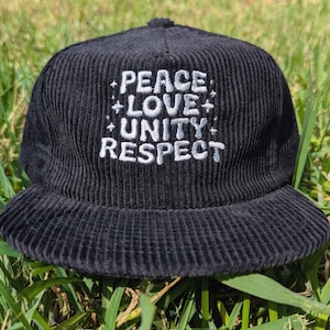 May include: Black corduroy baseball cap with the words "PEACE LOVE UNITY RESPECT" embroidered in white. The cap has a flat brim and is set against a background of green grass.