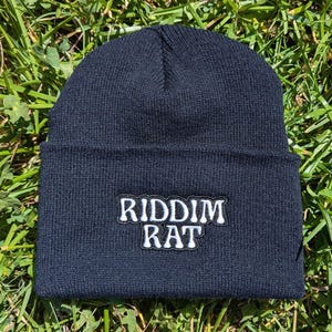 May include: A navy blue beanie with a white embroidered patch that reads "RIDDIM RAT". The hat is made of a knitted material and is displayed on a bed of green grass.