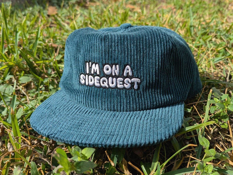 May include: Teal corduroy baseball cap with the embroidered text "I'M ON A SIDEQUEST" in white. The hat has a curved brim and a structured crown. The cap is sitting on a bed of green grass.