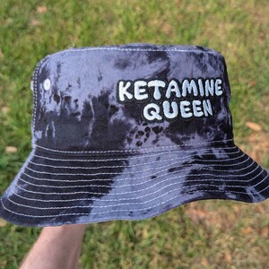 May include: A black and blue tie-dye bucket hat with the text "Ketamine Queen" embroidered in white on the front.