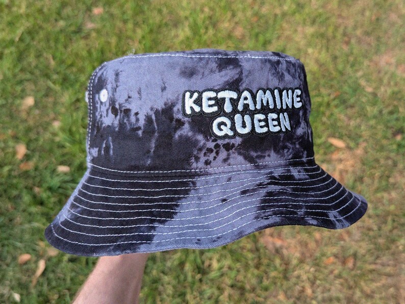 May include: A black and blue tie-dye bucket hat with the text "Ketamine Queen" embroidered in white on the front.