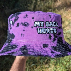 May include: A purple and black tie-dye bucket hat featuring the embroidered text MY BACK HURTS in white with a black outline. The hat has a flat top, a downward-sloping brim, and visible stitching details throughout the fabric construction.