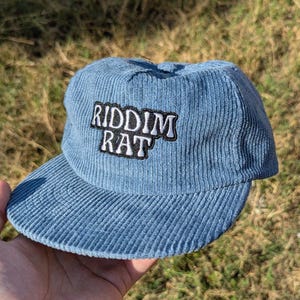 May include: A light blue corduroy cap with a curved brim. The cap features the text "RIDDIM RAT" in white letters with a black border. The hat is a casual accessory.