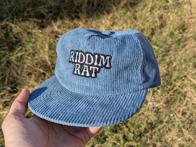 May include: A light blue corduroy cap with a curved brim. The cap features the text "RIDDIM RAT" in white letters with a black border. The hat is a casual accessory.
