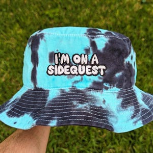 May include: A blue and black tie-dye bucket hat with the text "I'm on a Sidequest" embroidered in white.