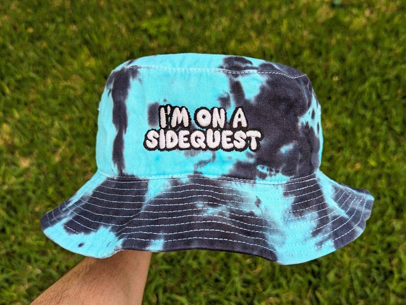 May include: A blue and black tie-dye bucket hat with the text "I'm on a Sidequest" embroidered in white.