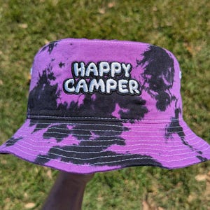 May include: A purple tie-dye bucket hat with the embroidered text "HAPPY CAMPER" in white. The hat has a black and purple pattern and a structured brim. The hat is a casual accessory.