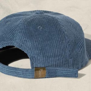 May include: A blue corduroy baseball cap with a curved brim and adjustable strap. The cap features a small, rectangular bronze-colored tag on the back. The cap is made of a soft, ribbed fabric.