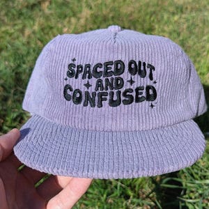 May include: A lavender corduroy baseball cap with the embroidered text "SPACED OUT and CONFUSED" in black. The cap has a curved brim and a structured crown. The background is green grass.