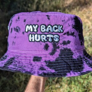 May include: A purple and black tie-dye bucket hat featuring the embroidered text MY BACK HURTS in white letters with a dark outline. The hat has a wide, downward-sloping brim and a flat crown, constructed from fabric with a visible stitched pattern.
