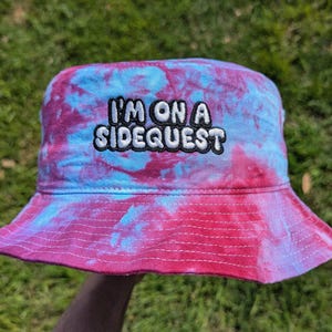 May include: A pink and blue tie-dye bucket hat with the text "I'm on a Sidequest" embroidered in black.