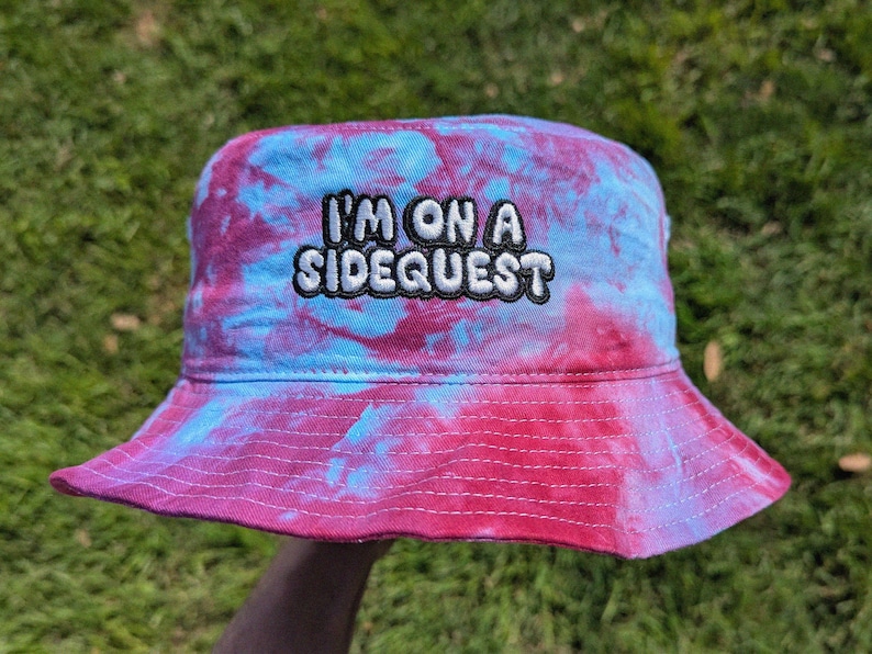 May include: A pink and blue tie-dye bucket hat with the text "I'm on a Sidequest" embroidered in black.