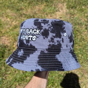 May include: A bucket hat featuring a black and grey tie-dye pattern. The front of the hat displays the embroidered white text "MY BACK HURTS" in a bold, rounded font. The hat has a downward-sloping brim and visible white stitching details.