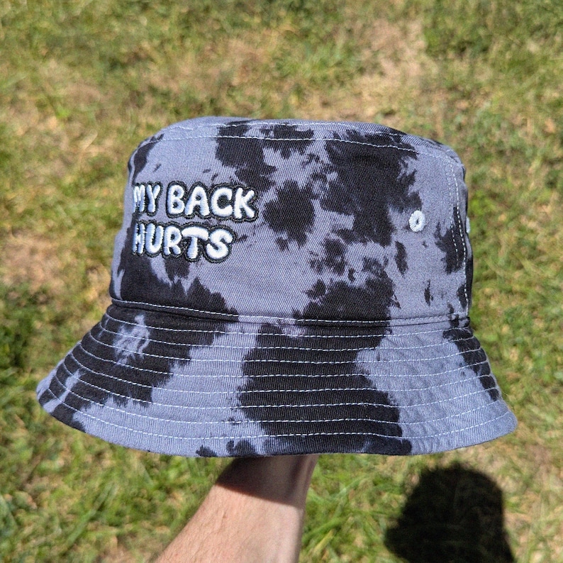 May include: A bucket hat featuring a black and grey tie-dye pattern. The front of the hat displays the embroidered white text "MY BACK HURTS" in a bold, rounded font. The hat has a downward-sloping brim and visible white stitching details.