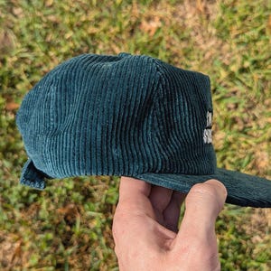 May include: Teal corduroy baseball cap with a flat brim. The cap has white embroidered text on the side. The hat is held up against a background of green grass.