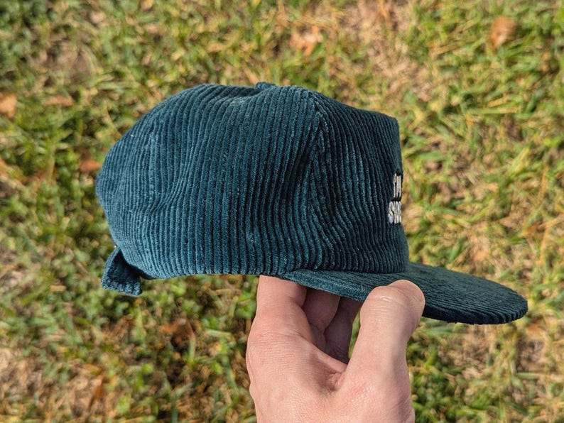 May include: Teal corduroy baseball cap with a flat brim. The cap has white embroidered text on the side. The hat is held up against a background of green grass.