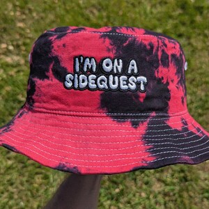 May include: Red and black tie-dye bucket hat with the text "I'm on a Sidequest" embroidered in white.