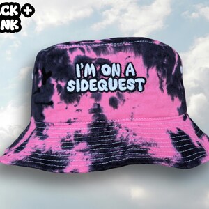 May include: Black and pink tie-dye bucket hat with the text "I'm on a Sidequest" embroidered in white.