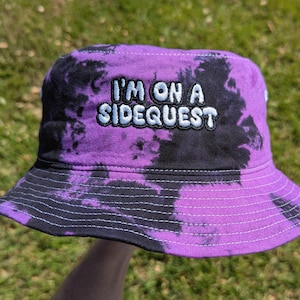 May include: Purple and black tie-dye bucket hat with the text "I'm on a Sidequest" embroidered in white.