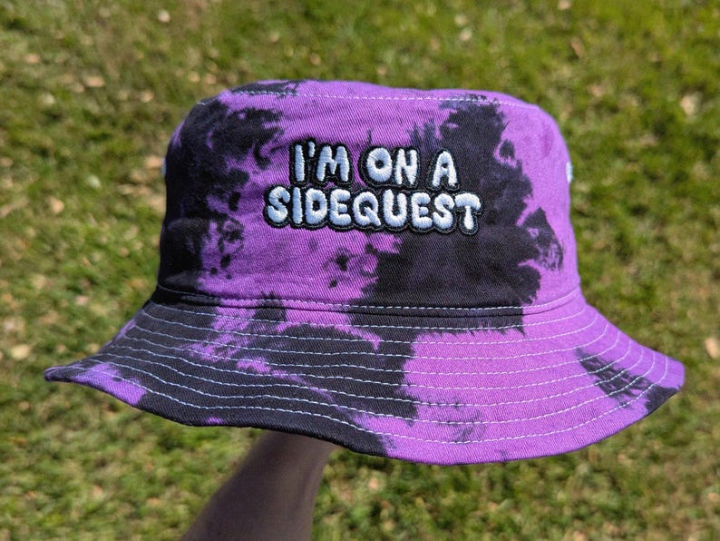 May include: Purple and black tie-dye bucket hat with the text "I'm on a Sidequest" embroidered in white.