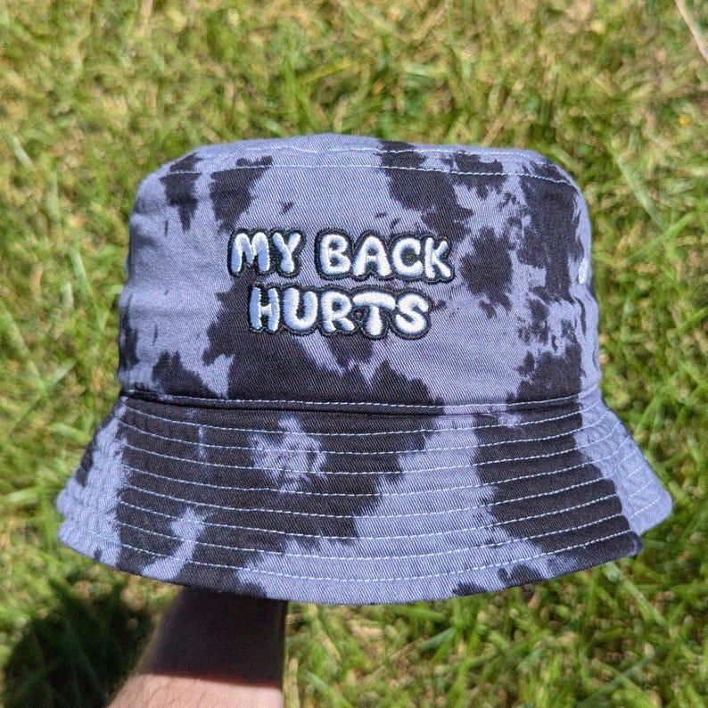 May include: A bucket hat featuring a grey and black tie-dye pattern. The front of the hat displays the embroidered text MY BACK HURTS in white, bubbly, outlined letters. The hat has a downward-sloping brim and visible stitching along the crown.