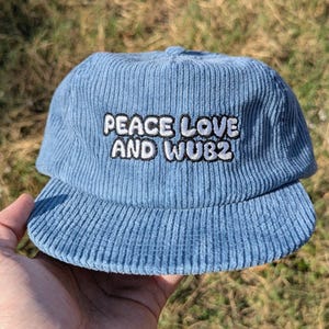 May include: A light blue corduroy baseball cap with the embroidered text "PEACE LOVE AND WUB2" in white with a black outline. The cap has a curved brim and a rounded crown.
