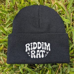 May include: A black beanie with the white text "RIDDIM RAT" embroidered on the front. The text is stylized with a playful font and star accents. The beanie is made of a soft, knitted material.