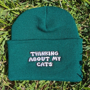 May include: A dark teal beanie with the embroidered text "THINKING ABOUT MY CATS" in white, outlined in black. The beanie is made of a soft, knitted material and is displayed on a bed of green grass.