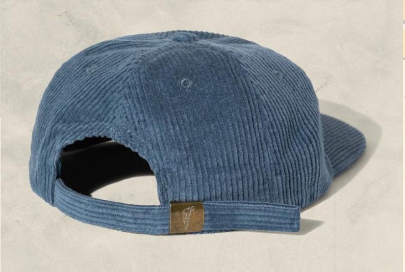 May include: A blue corduroy baseball cap with a curved brim and adjustable strap. The cap features a small, rectangular bronze-colored tag on the back. The cap is made of a soft, ribbed fabric.