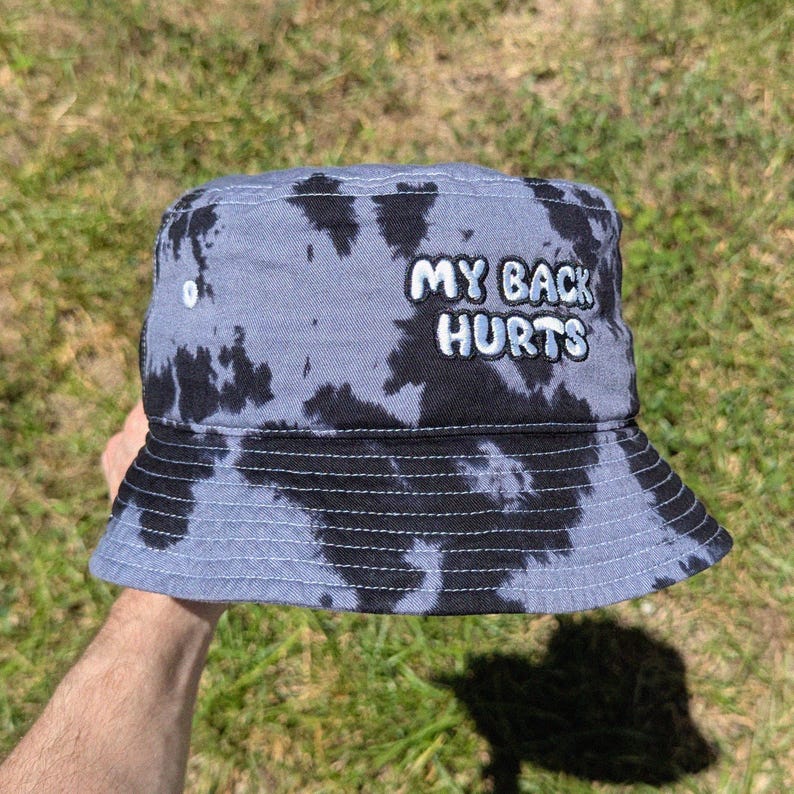 May include: A grey and black tie-dye bucket hat featuring the embroidered white text MY BACK HURTS on the front. The hat has a flat top, a downward-sloping brim with horizontal stitching, and a small white ventilation eyelet on the side panel.