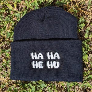 May include: Black beanie with the text "HA HA HE HU" in white, outlined in black. The beanie is made of a soft, knitted material and is suitable for cold weather. The hat is laying on a bed of green grass.