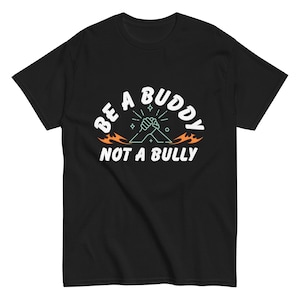 Puede incluir: Camiseta negra con texto blanco que dice "Be a Buddy Not a Bully". El texto está en un círculo con llamas a cada lado. Hay dos manos juntas en el centro del círculo.