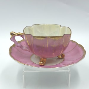 May include: A pink teacup and saucer set with gold trim. The teacup has a unique shape with a scalloped rim and sits on three small gold feet. The saucer is also pink with a scalloped edge, creating an elegant vintage aesthetic.